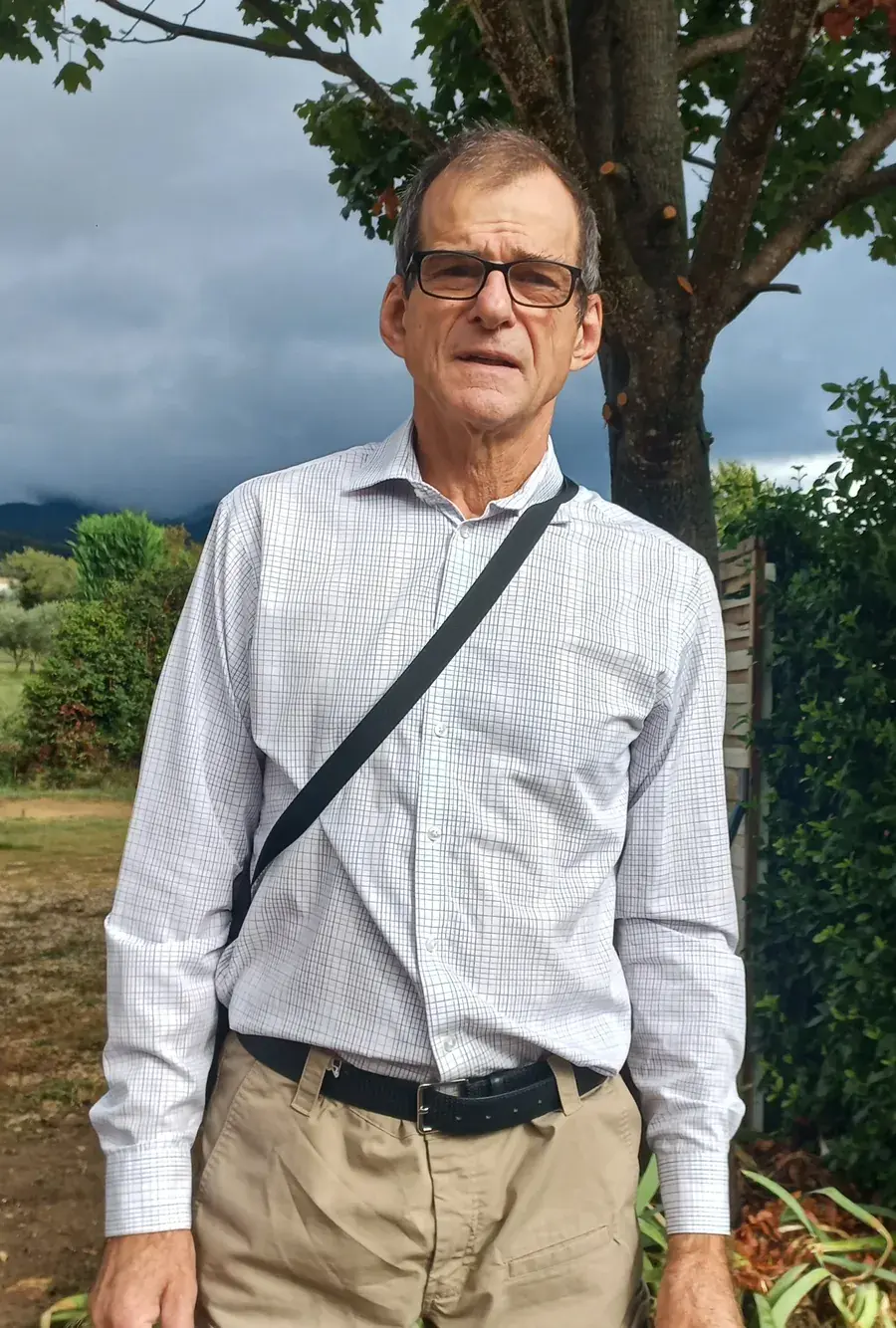 Dominique Beord, chauffeur VTC, en chemise devant un arbre, le Luberon sous les nuages derrière lui